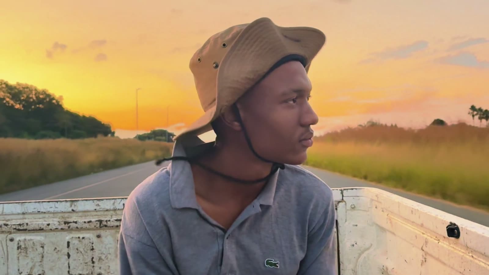 A young man in a bush hat rides on the back of a bakkie, an orange sky behind him.