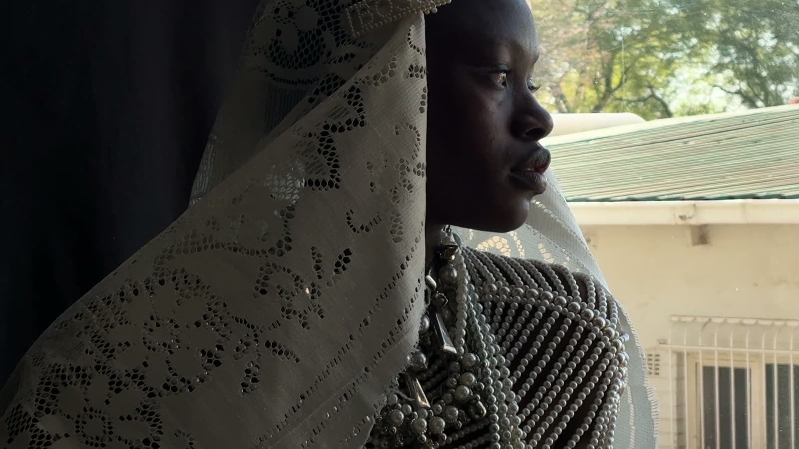 A woman in a lace veil and pearl bodice looks out of a window, side-lit by daylight.