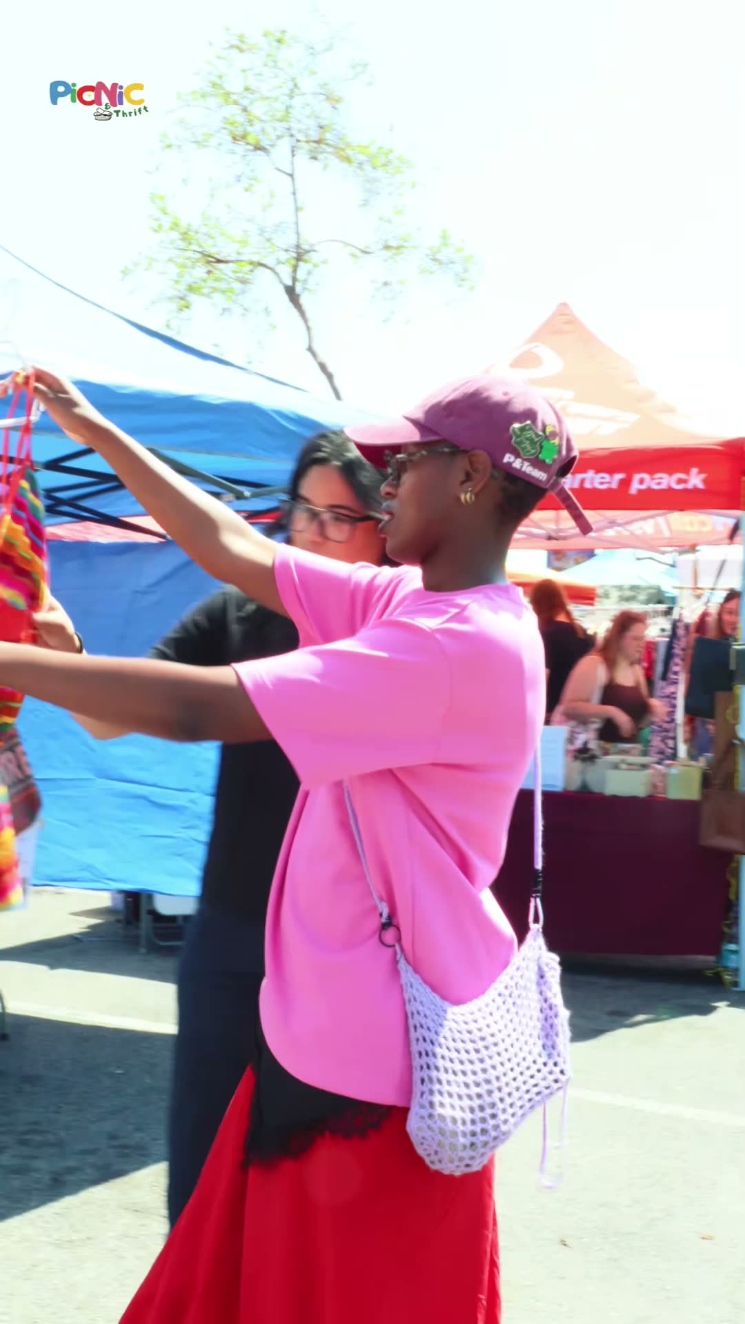 A shopper in pink dances beside the clothing rails at a sunny outdoor market.