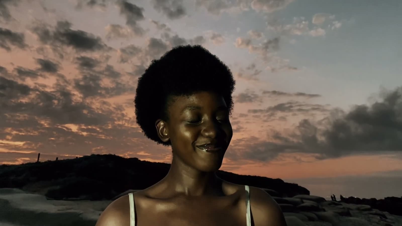 A woman smiles softly against a pink and grey sunset sky on the beach.